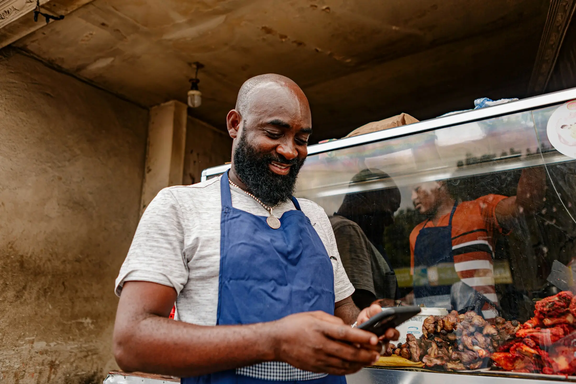 Food stall owner in a blue apron checking orders on his phone behind a counter of freshly grilled food