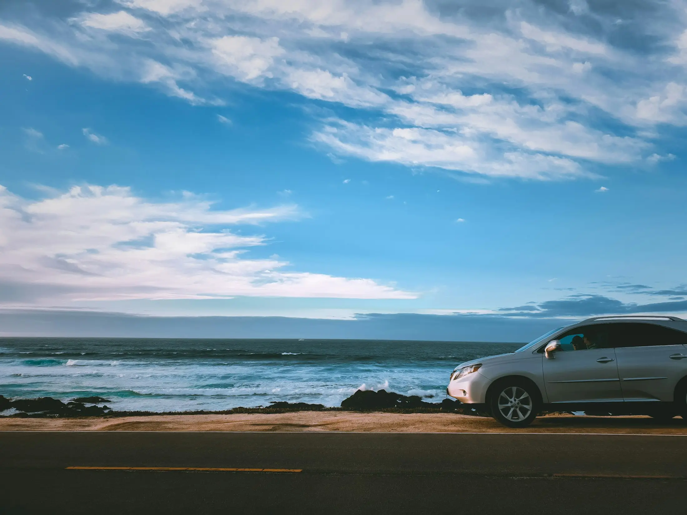 Silver SUV parked on a coastal road with the ocean and a wide blue sky behind it