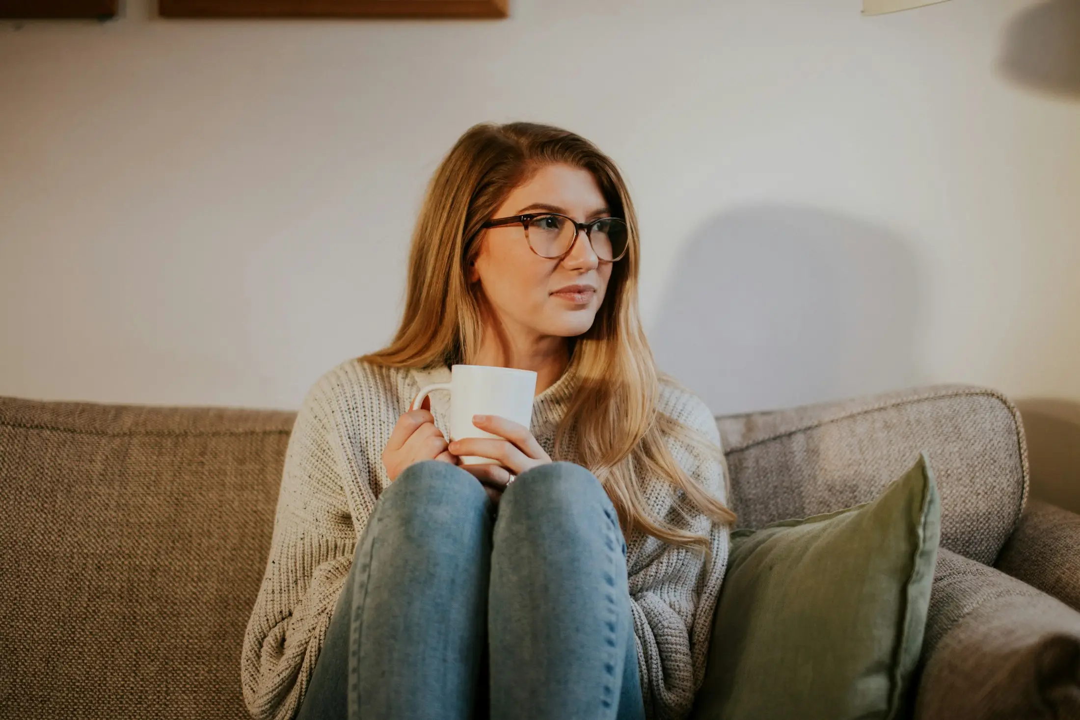 Woman in a cosy knit jumper curled up on a sofa with a cup of tea, looking calm