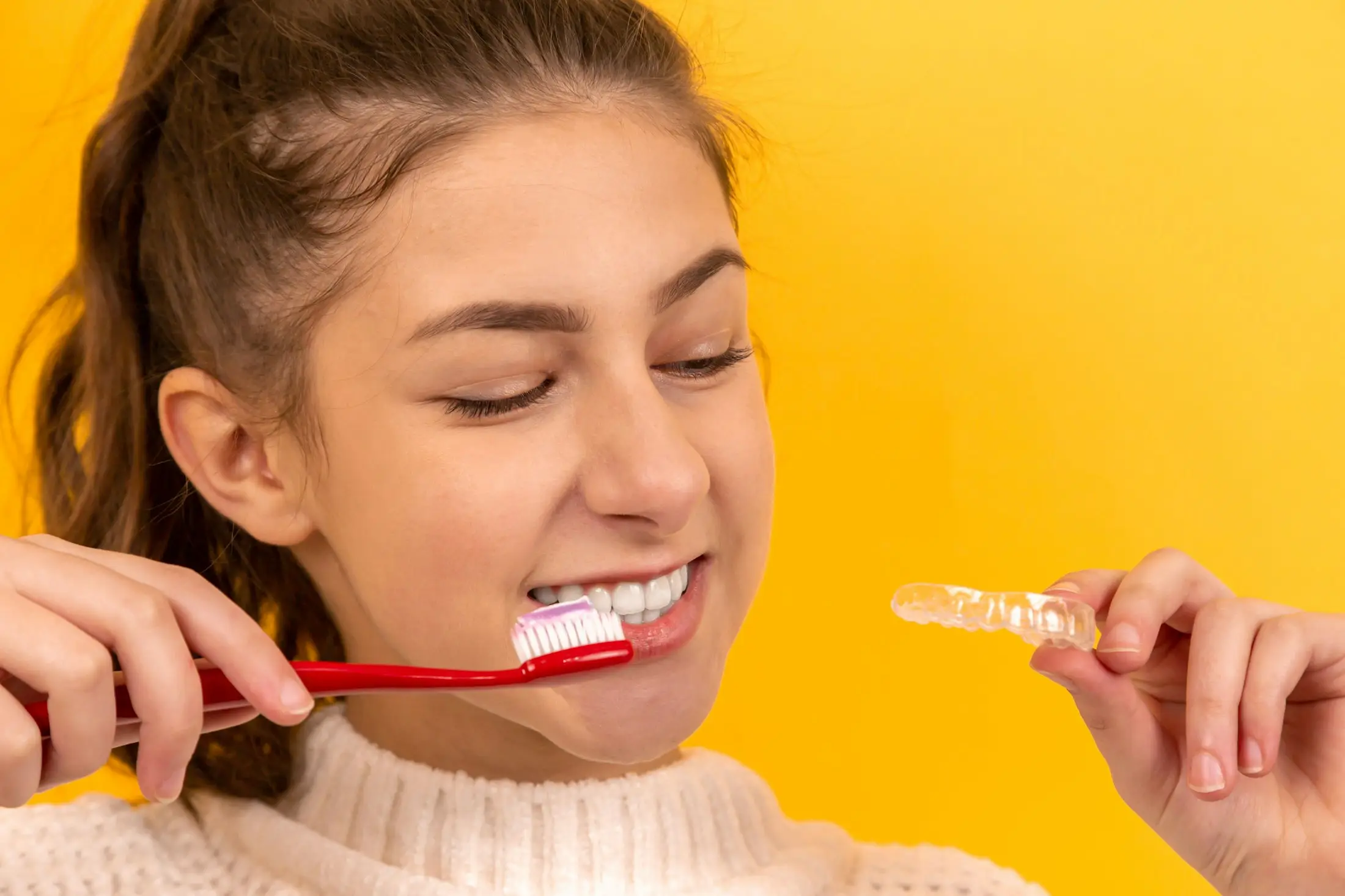 Young woman smiling as she brushes her teeth and holds up a clear aligner against a yellow background