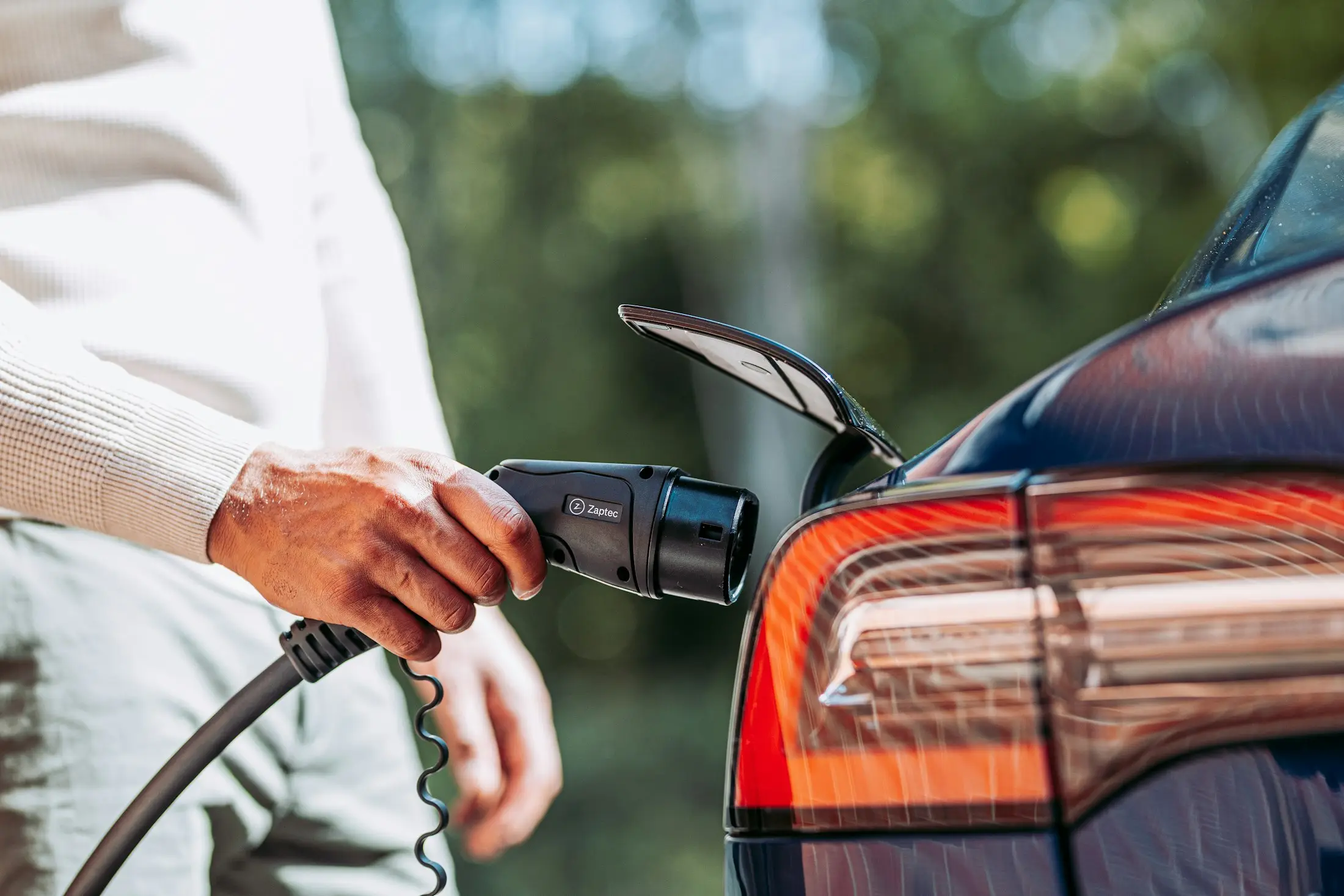 A hand holding a charging connector up to the open charge port of an electric car
