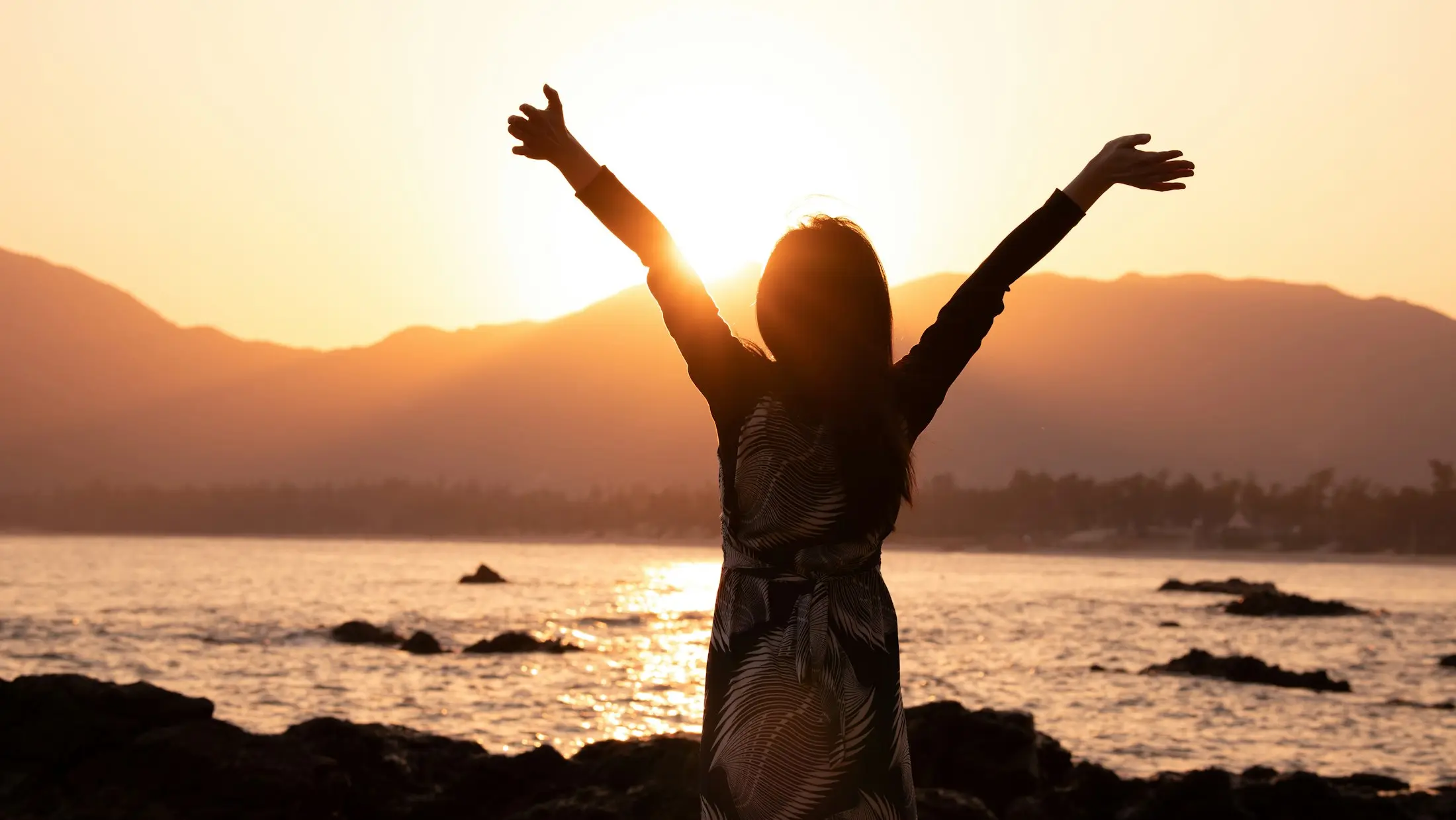 Woman silhouetted against a golden sunset over the sea, arms raised in celebration