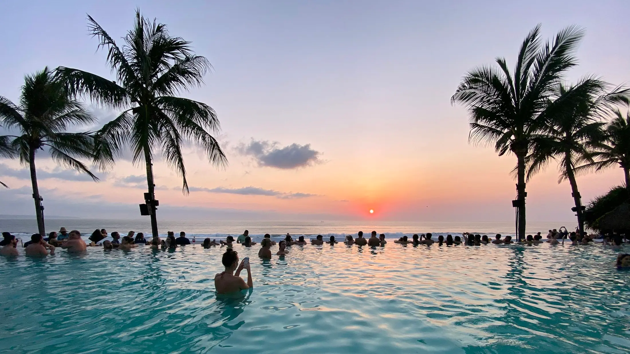 Swimmers in an infinity pool at sunset framed by palm trees