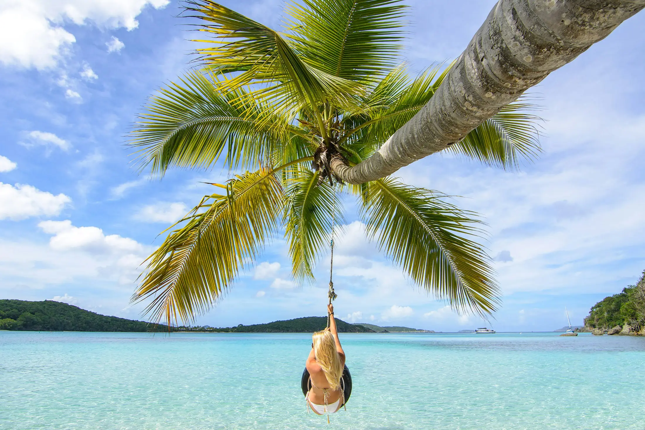 Traveller on a rope swing hanging from a leaning palm over clear water