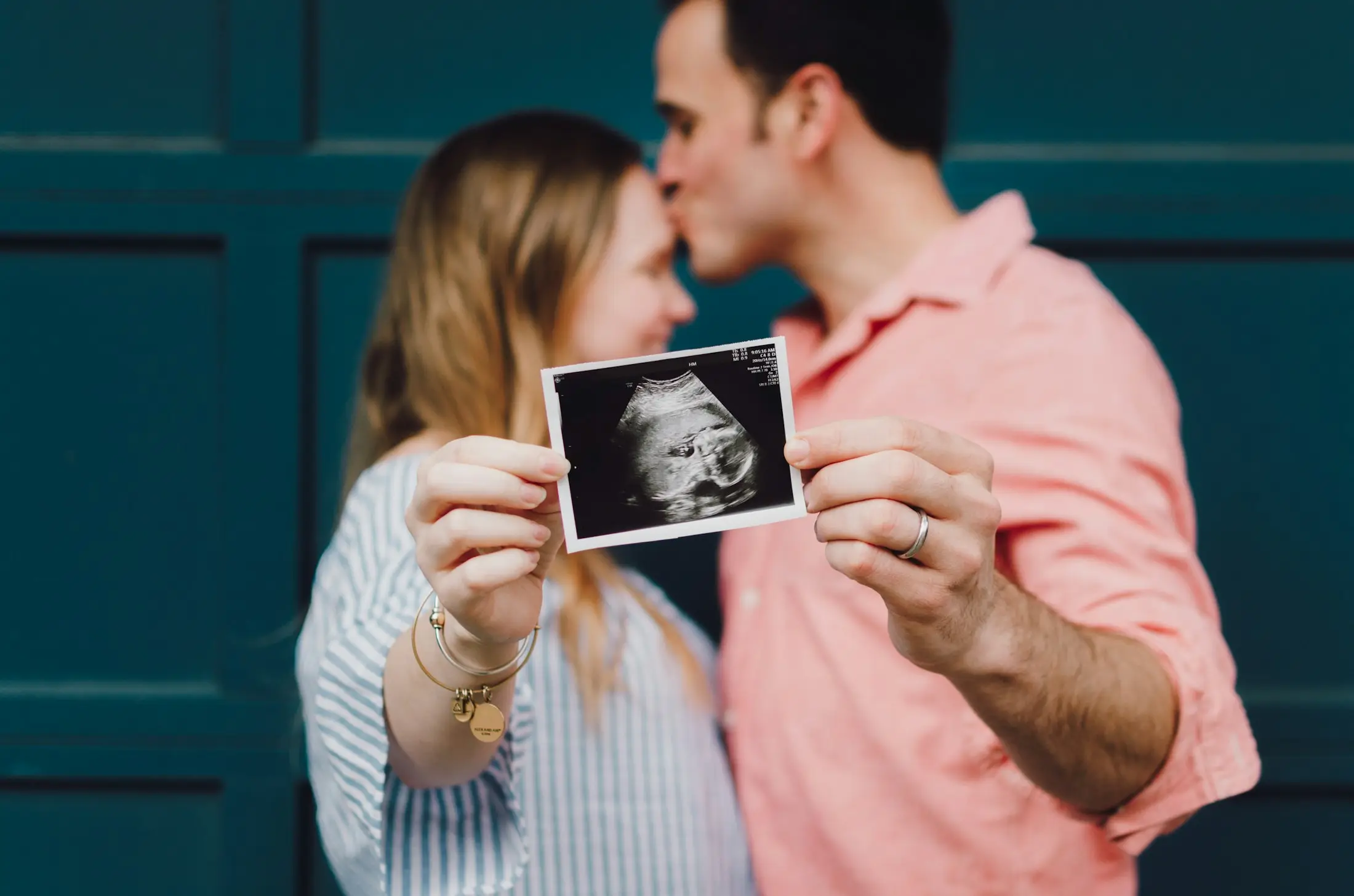 Couple holding up an ultrasound photo as the man gently kisses the woman’s forehead