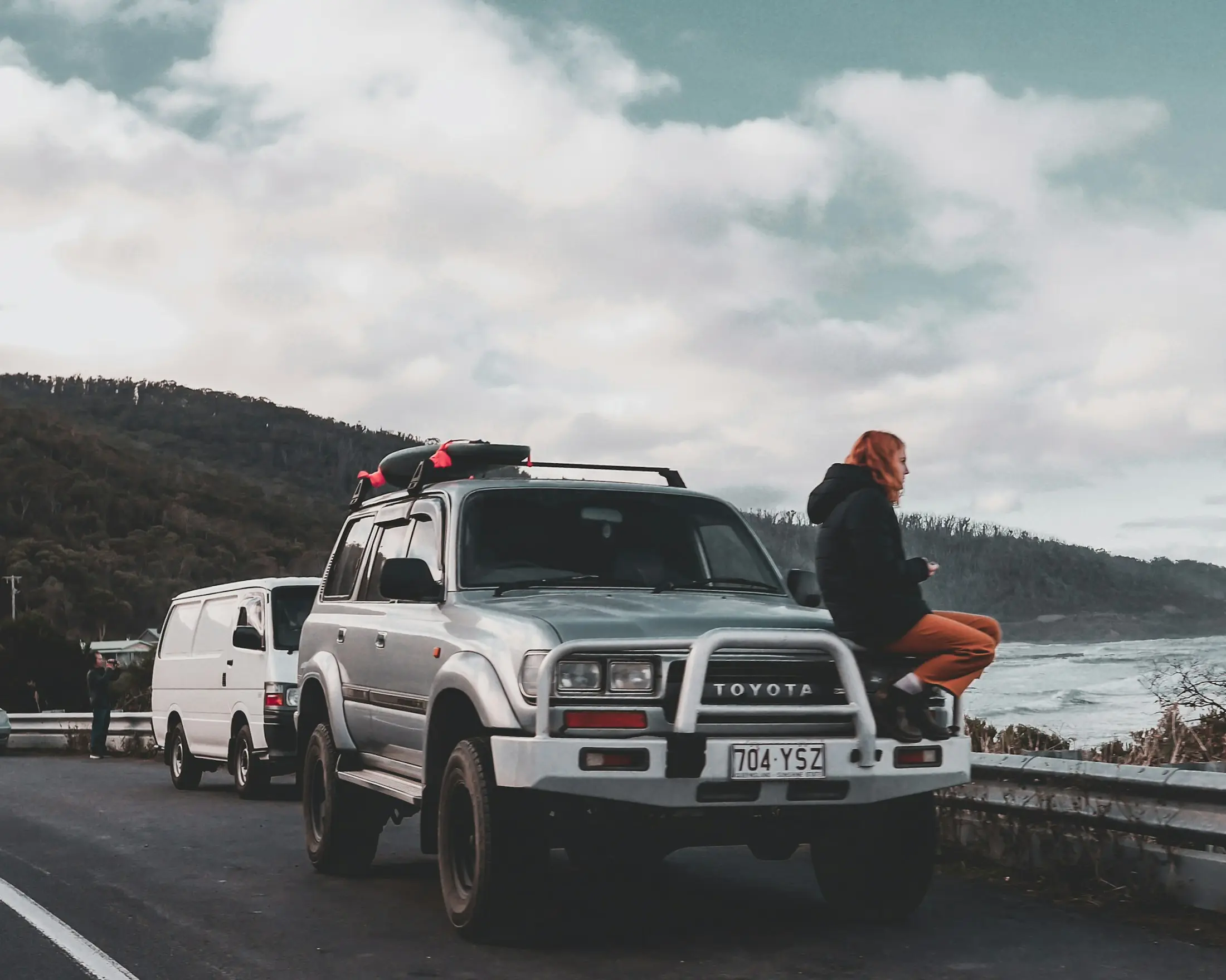 Traveller sitting on the bonnet of a 4WD with a roof pod, parked on a coastal road beside a campervan