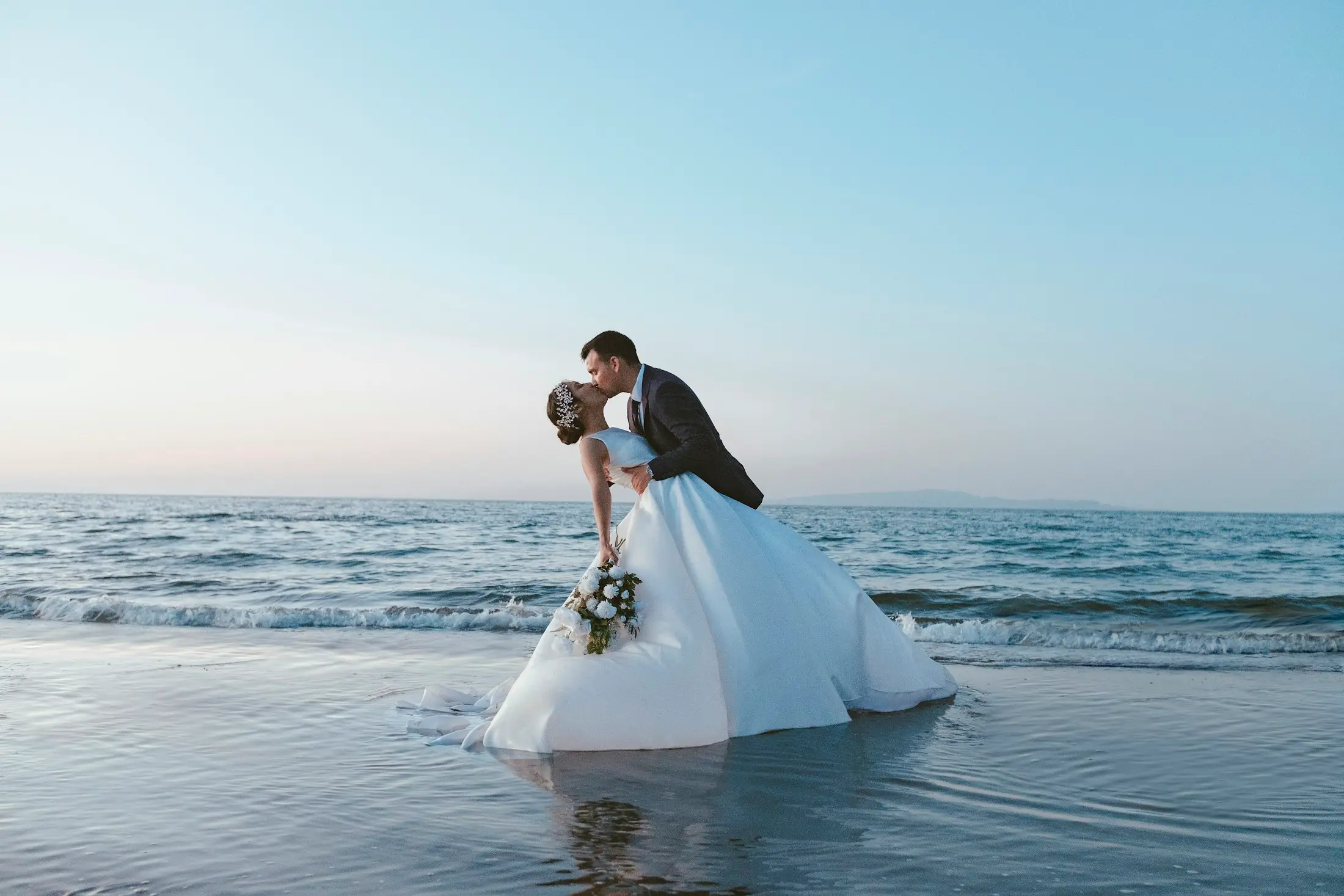 Bride and groom kissing at the water’s edge on a beach, her white gown trailing in the shallows